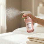 Hand spraying LEAFI Aroma Secret Flower room spray mist over fresh white bed linen in a bright bedroom